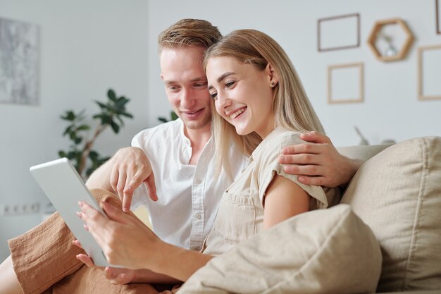 Fröhlicher junger liebevoller Mann, der seine hübsche lächelnde Frau umarmt und auf den Bildschirm des digitalen Tablets zeigt, während er durch den Online-Shop scrollt