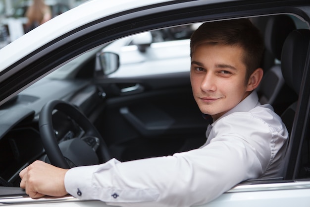 Fröhlicher junger männlicher Fahrer, der in die Kamera lächelt und in einem neuen Auto im Autohaus sitzt