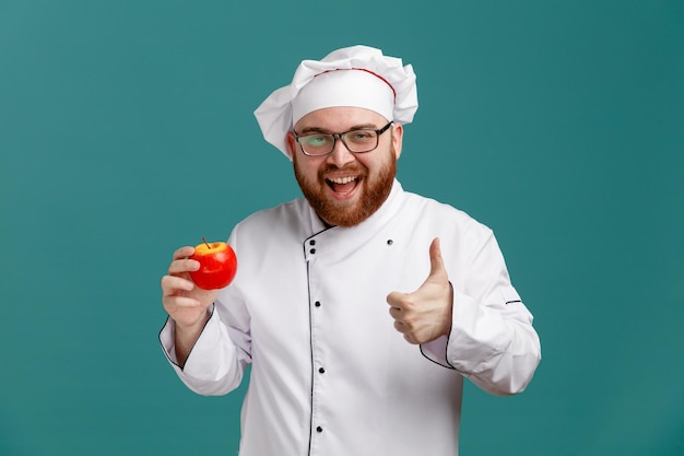 Fröhlicher junger männlicher Koch mit einheitlicher Brille und Mütze mit Apfel, der in die Kamera schaut und den Daumen isoliert auf blauem Hintergrund zeigt