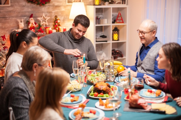 Fröhlicher junger Mann, der beim Weihnachtsessen mit der Familie eine Flasche Wein öffnet.