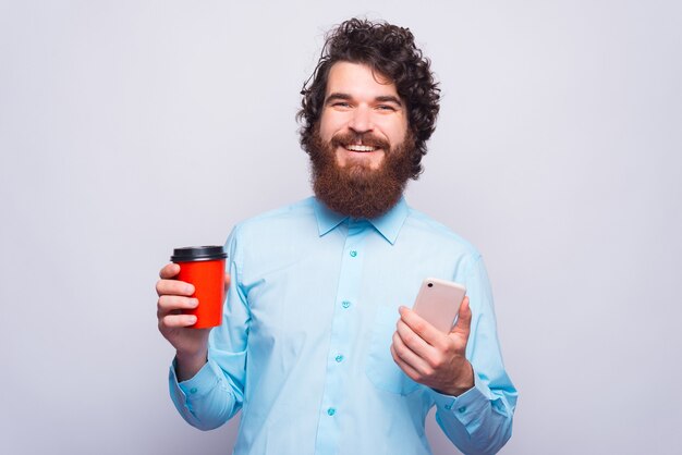 Fröhlicher junger Mann, der lächelt und in die Kamera schaut, hält eine Tasse mit heißem Getränk und einem Telefon