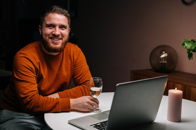 Fröhlicher junger Mann, der sich mit einem Laptop aus der Ferne verabredet, ein Glas mit Wein in der Hand hält und per Videoanruf anstößt. Verliebtes Paar chattet bei einem Remote-Date.