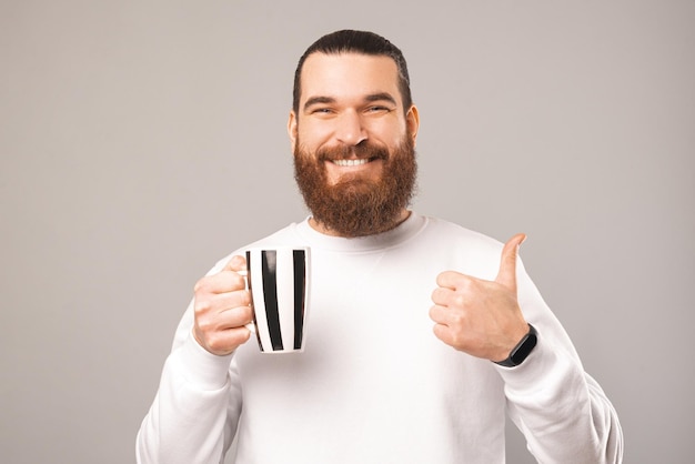 Fröhlicher junger Mann hält eine schwarz-weiße Tasse, während er den Daumen nach oben zeigt