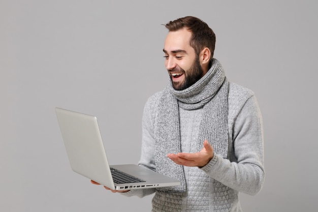 Fröhlicher junger Mann im grauen Pullover, Schal isoliert auf grauem Hintergrund. Gesunder Lebensstil kranke Krankheit Online-Behandlungsberatung, Konzept der kalten Jahreszeit. Mock-up-Kopienbereich. Halten Sie einen Laptop-PC-Computer.