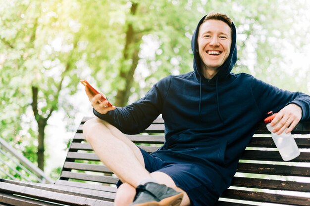 Fröhlicher junger Mann im Hoodie, der auf der Bank sitzt und sich nach dem Sporttraining mit dem Handy entspannt