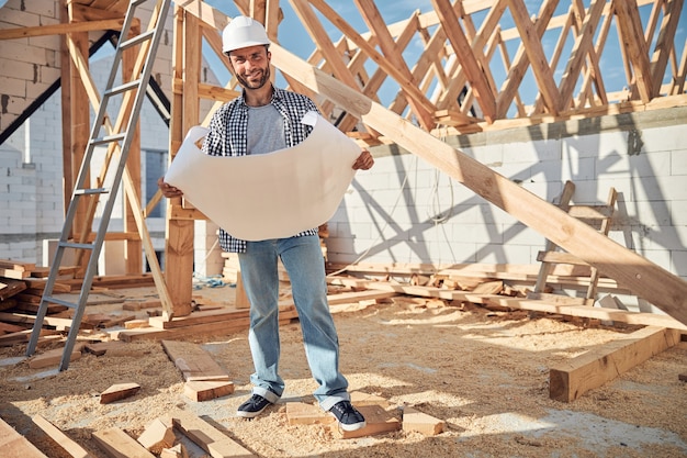 Fröhlicher junger Mann in einem Bauarbeiterhelm, der einen Bauplan hält