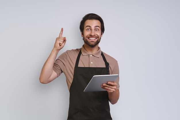 Fröhlicher junger Mann in Schürze, der sein digitales Tablet zeigt und gestikuliert, während er gegen grauen B...