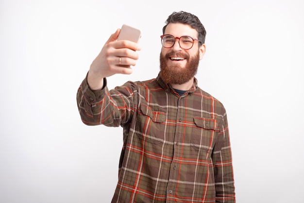 Fröhlicher junger Mann macht ein Selfie mit seinem Telefon auf weißem Hintergrund.