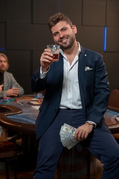 Fröhlicher junger Mann mit Banknoten, der ein Glas Whiskey für ein erfolgreiches Pokerspiel hebt