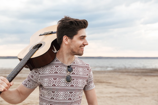 Fröhlicher junger Mann mit Gitarre, am Strand stehend