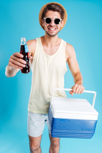 Fröhlicher junger Mann mit Hut und Sonnenbrille, der Ihnen eine Kühltasche und eine Flasche Soda über der blauen Wand gibt