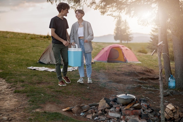 Fröhlicher junger Mann und Frau, die mit tragbarem Kühlschrank am Campingplatz stehen und lachen