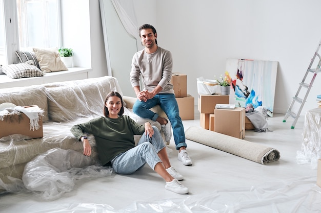 Fröhlicher junger Mann und Frau in Jeans und Pullovern, die nach dem Entfernen auf der Couch im Wohnzimmer ihres neuen Hauses oder ihrer neuen Wohnung sitzen