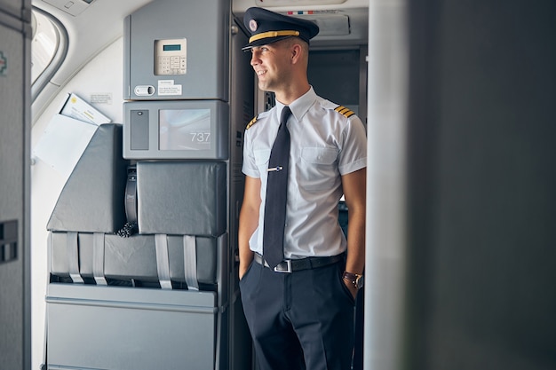 Fröhlicher junger Pilot mit Kapitänshut, der wegschaut und lächelt, während er die Hände in den Taschen hält