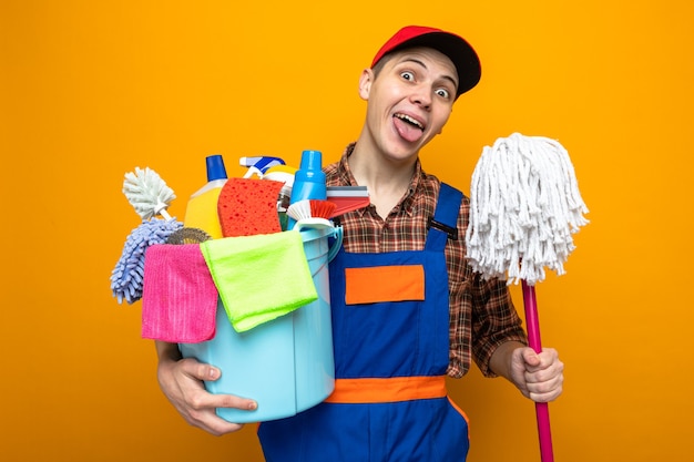 Fröhlicher junger Putzmann, der eine Uniform und eine Kappe trägt, die einen Eimer mit Reinigungswerkzeugen mit Mop hält