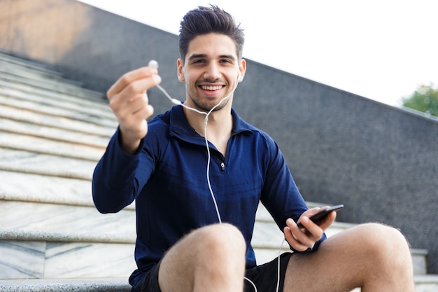 Fröhlicher junger Sportler, der Musik mit Kopfhörern hört, während draußen sitzt