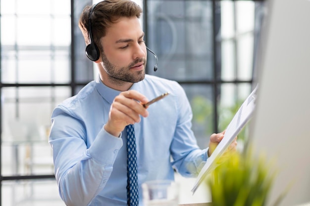 Fröhlicher junger Support-Telefon-Operator im Headset am Arbeitsplatz bei der Verwendung des Computers