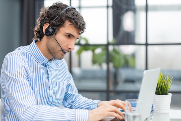 Fröhlicher junger Support-Telefon-Operator im Headset am Arbeitsplatz, während er den Computer-Help-Service und das Kundenberatungs-Call-Center-Konzept nutzt