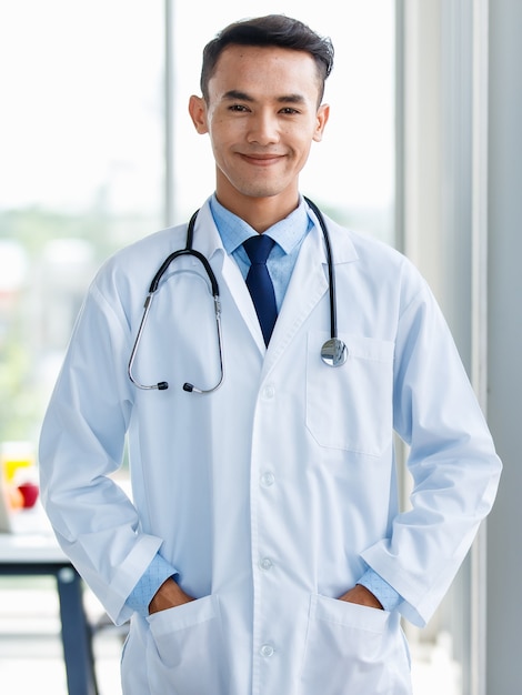 Fröhlicher junger und süßer asiatischer männlicher Arzt in Uniform und mit Stethoskop, der in der Klinik steht und mit positiver und selbstbewusster Geste posiert.