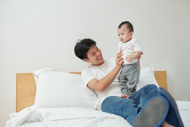 Fröhlicher junger vietnamesischer Mann, der auf Bett sitzt und mit entzückender kleiner Tochter spielt