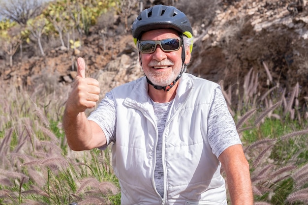 Fröhlicher kaukasischer älterer Mann bei Landausflügen mit Sporthelm und Sonnenbrille, die ein positives Zeichen in Ordnung gestikulieren