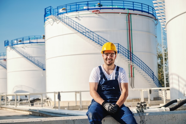 Fröhlicher kaukasischer Arbeiter insgesamt und mit Helm auf dem Kopf, der draußen sitzt.