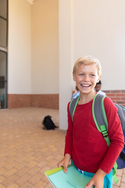 Fröhlicher kaukasischer Grundschüler mit Büchern, der auf dem Schulgelände wegblickt