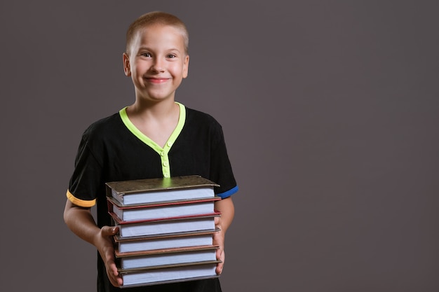 Fröhlicher kaukasischer Junge in einem schwarzen T-Shirt hält einen Stapel Bücher auf grauem Hintergrund. Bildungskonzept