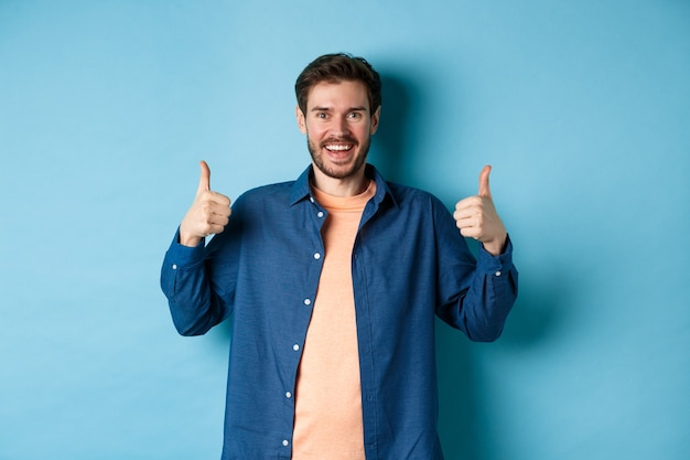 Fröhlicher kaukasischer Kerl im Freizeithemd, der Daumen oben in der Zustimmung zeigt, lächelt und etwas Gutes lobt, das auf blauem Hintergrund steht.