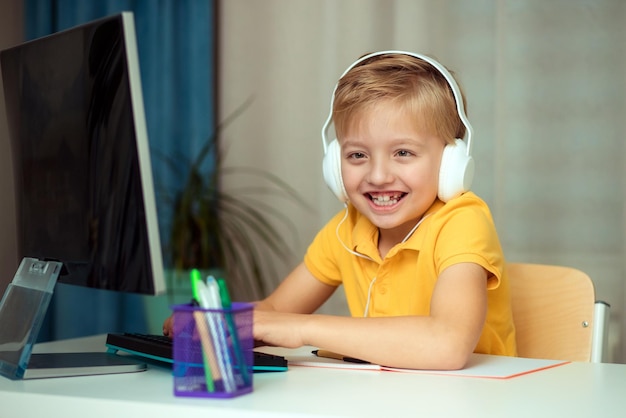 Fröhlicher Kinderjunge in Kopfhörern beim Online-Lernen zu Hause bei Hausaufgaben