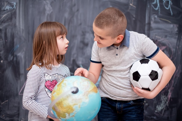 Fröhlicher Kinderjunge mit Fußball und kleinem Mädchen, das mit dem Erdball etwas über die Welt lernt, während er vor einer schwarzen Tafel steht