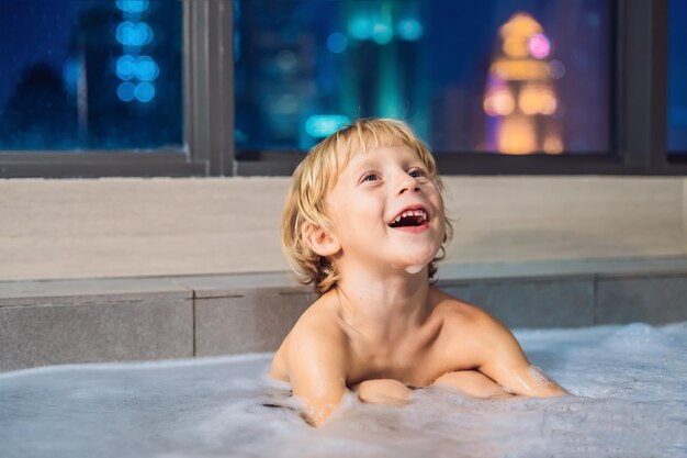 Fröhlicher kleiner Junge, der abends in der Badewanne sitzt, bevor er auf dem Hintergrund eines Fensters mit Blick auf die abendliche Stadt schlafen geht. Porträt eines Babys, das in einer Badewanne voller Schaum in der Nähe des Fensters badet