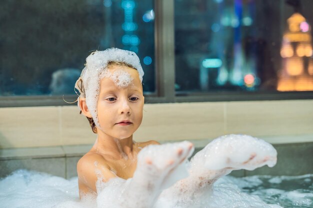 Fröhlicher kleiner Junge, der abends in der Badewanne sitzt, bevor er auf dem Hintergrund eines Fensters mit Blick auf die abendliche Stadt schlafen geht. Porträt eines Babys, das in einer Badewanne voller Schaum in der Nähe des Fensters badet