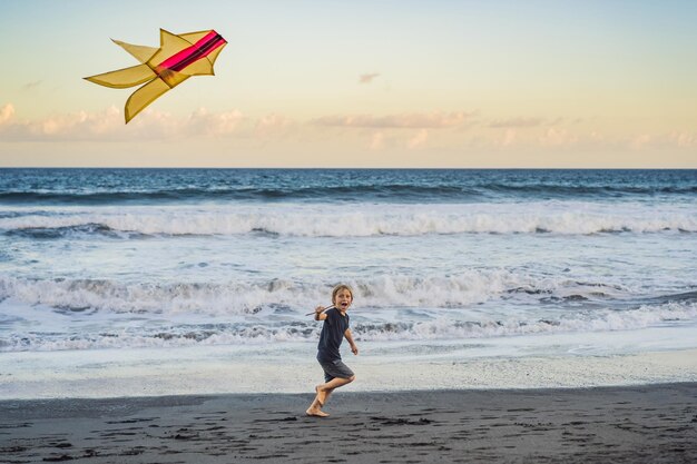 Fröhlicher kleiner Junge, der Drachen am Strand bei Sonnenuntergang fliegt