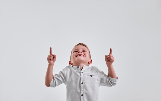 Foto fröhlicher kleiner junge, der mit beiden händen über grauem hintergrund schaut und nach oben zeigt und lächelt.