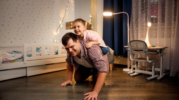 Fröhlicher kleiner Junge, der nachts auf seinem Vater huckepack reitet. Konzept des Kindes, das mit Eltern und Familie spielt, die nachts Zeit zusammen haben.