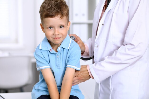 Fröhlicher kleiner Junge, der Spaß hat, während er vom Arzt mit Stethoskop untersucht wird Krankenversicherung und Hilfekonzept