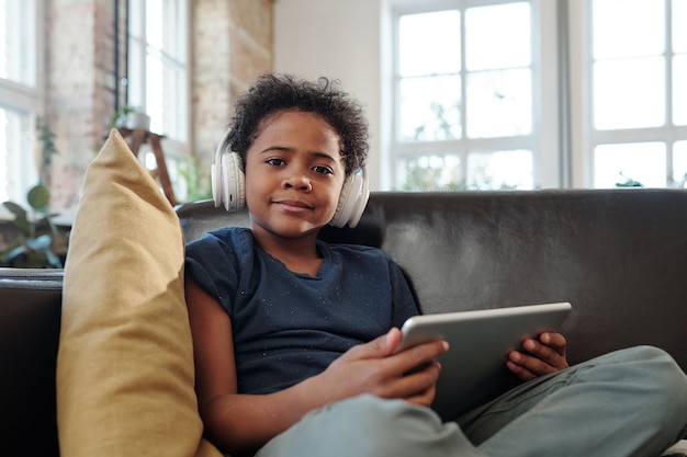 Fröhlicher kleiner Junge mit Kopfhörern und Tablet, der Sie ansieht, während er auf der Couch vor der Kamera sitzt und Online-Videos in der häuslichen Umgebung anschaut