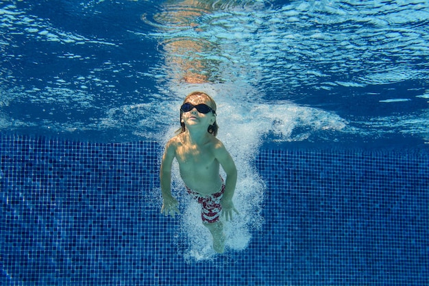 Fröhlicher kleiner Junge mit Schutzbrille und Shorts schwimmt unter Wasser im Pool mit Spritzern und Blasen