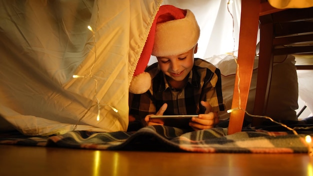 Fröhlicher kleiner Junge mit Weihnachtsmütze, der mit dem Smartphone im DIY-Kid39-Zelt liegt