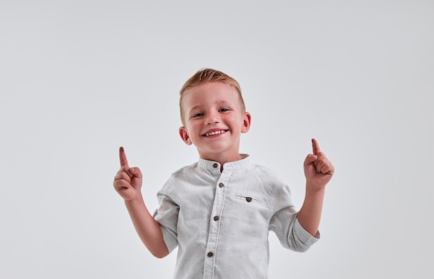 Foto fröhlicher kleiner junge zeigt mit beiden händen auf grauem hintergrund und lächelt.