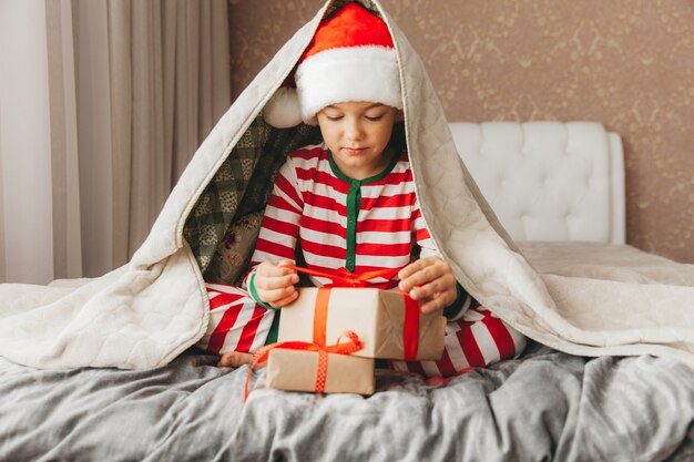 Fröhlicher lächelnder fröhlicher Kinderjunge im Weihnachtsmann-Hut, der ein Weihnachtsgeschenk in seinen Händen hält und auf dem Bett sitzt.