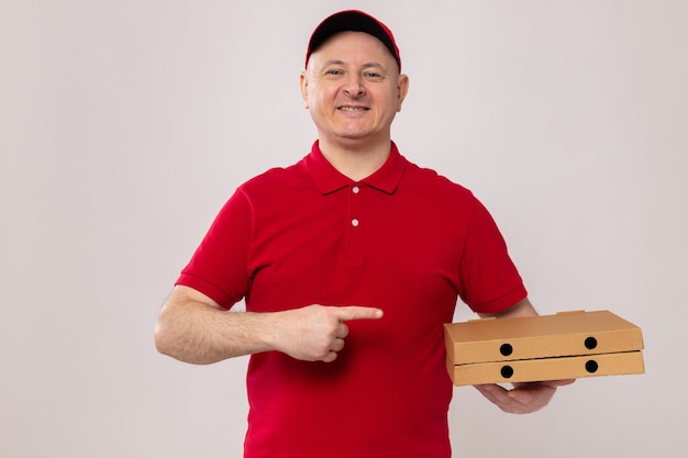 Fröhlicher Liefermann in roter Uniform und Mütze, der Pizzakartons hält und mit dem Zeigefinger auf sie zeigt, fröhlich lächelnd auf weißem Hintergrund stehend