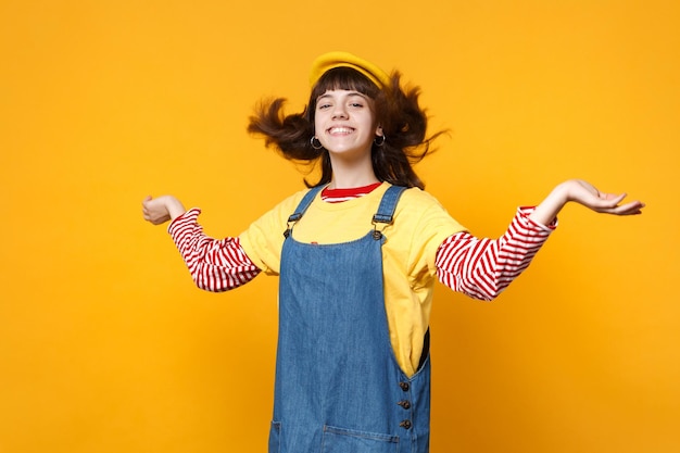 Fröhlicher Mädchen-Teenager im französischen Beret-Denim-Sommerkleid, das die Hände mit flatterndem Haar ausbreitet, isoliert auf gelbem Wandhintergrund im Studio. Menschen aufrichtige Emotionen, Lifestyle-Konzept. Mock-up-Kopienbereich.