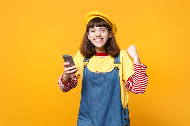 Fröhlicher Mädchen-Teenager in französischer Baskenmütze, Denim-Sommerkleid in der Hand und mit Handy, Gewinnergeste isoliert auf gelbem Hintergrund. Menschen aufrichtige Emotionen, Lifestyle-Konzept. Mock-up-Kopienbereich.