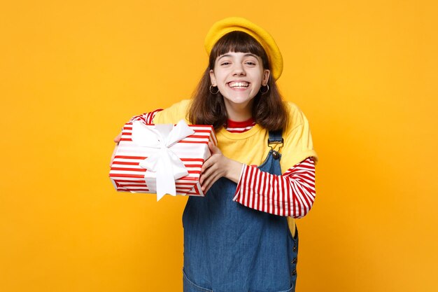 Fröhlicher Mädchen-Teenager in französischer Baskenmütze, Denim-Sommerkleid mit rot gestreifter Geschenkbox mit Geschenkband isoliert auf gelbem Wandhintergrund. Menschen aufrichtige Emotionen, Lifestyle-Geburtstagsurlaubskonzept.