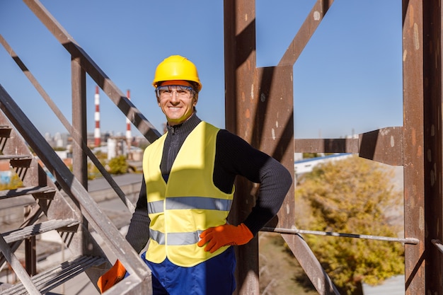 Fröhlicher männlicher Arbeiter, der in der Nähe der Leiter am Industriestandort steht