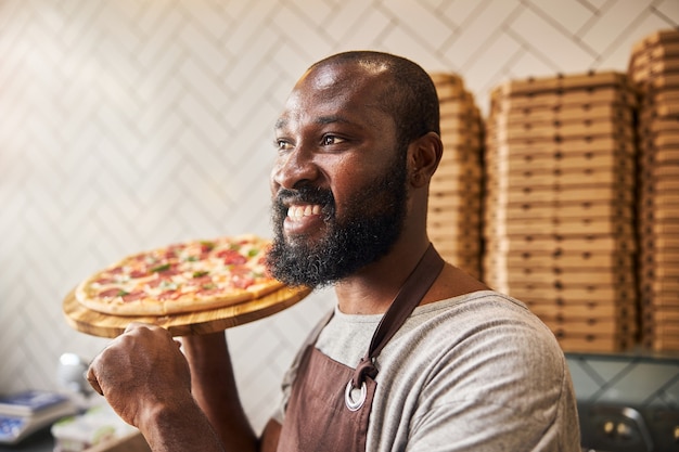Fröhlicher männlicher Arbeiter mit leckerer Pizza in den Händen, der wegschaut und lächelt