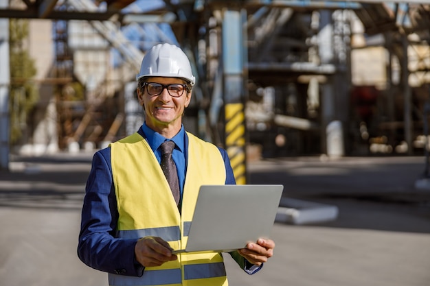 Fröhlicher männlicher Ingenieur mit Notebook im Freien in der Fabrik