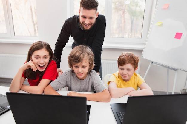 Fröhlicher männlicher Lehrer und Gruppe von Schülern, die Software auf Laptop testen, während sie an Projekt während des technischen Unterrichts im modernen Klassenzimmer in der Schule arbeiten